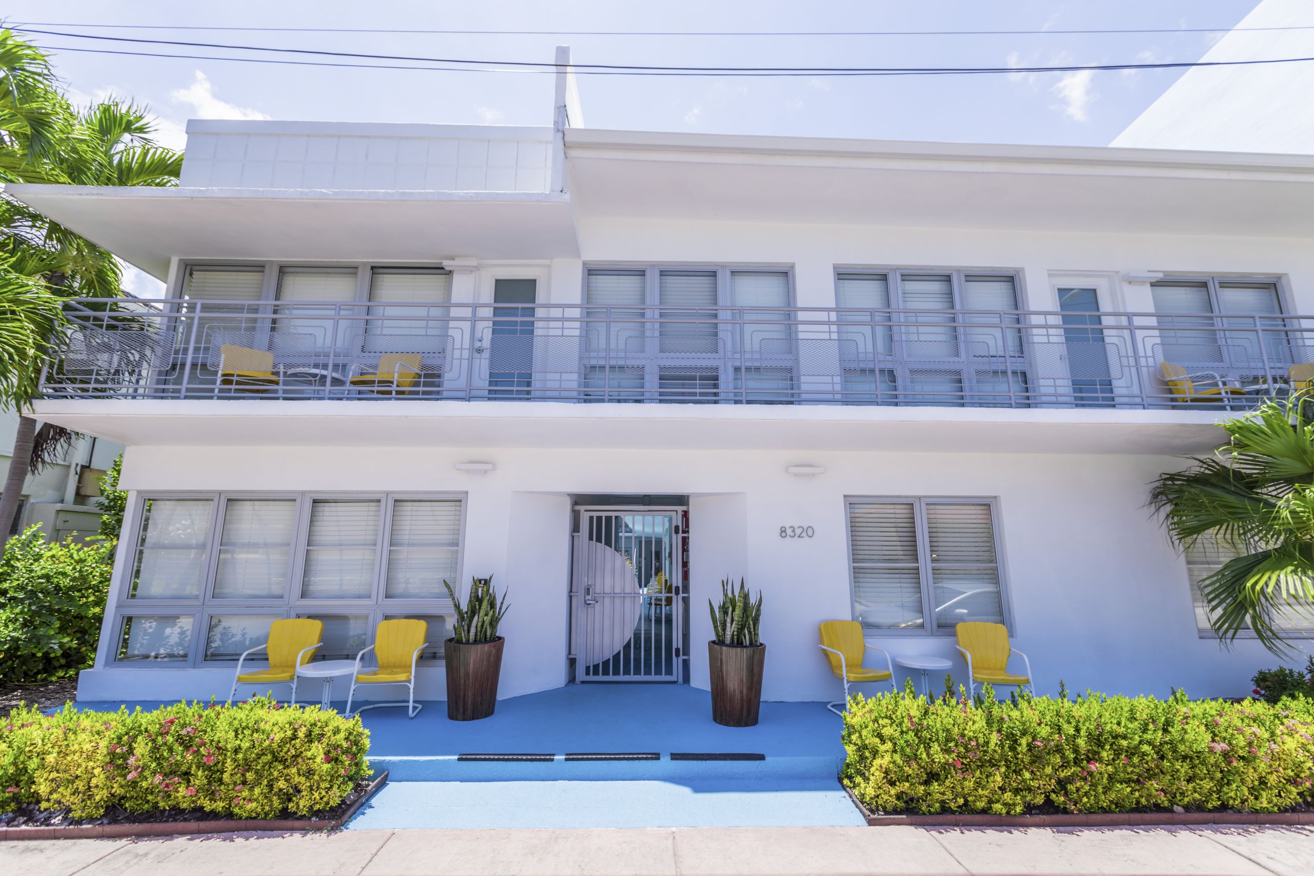 A white two-story building with balconies, yellow chairs, and potted plants on the ground floor.