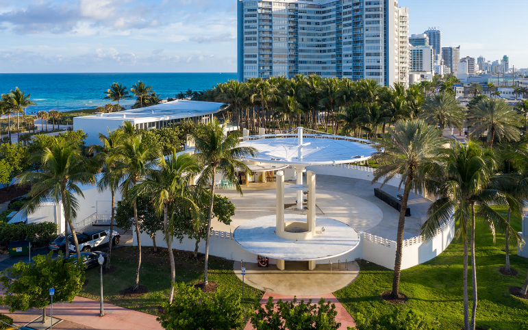 Aerial view of a modern white building with a circular structure surrounded by palm trees and overlooking the ocean and city skyline.